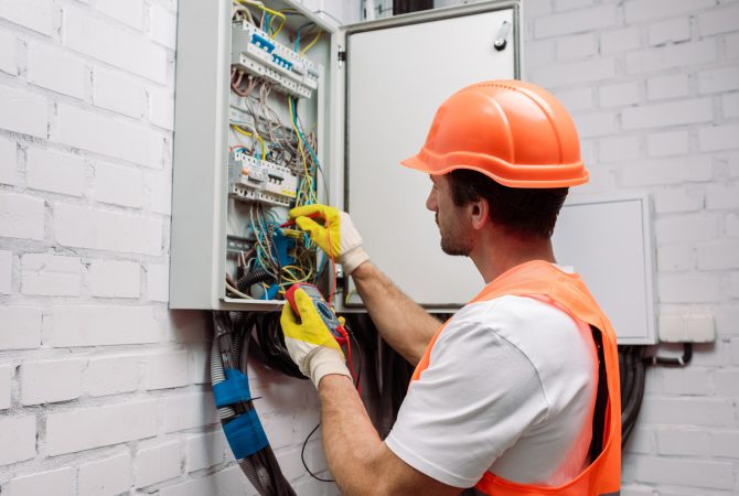 handsome-electrician-in-hardhat-and-gloves-holding-multimeter-near-electrical-distribution-box