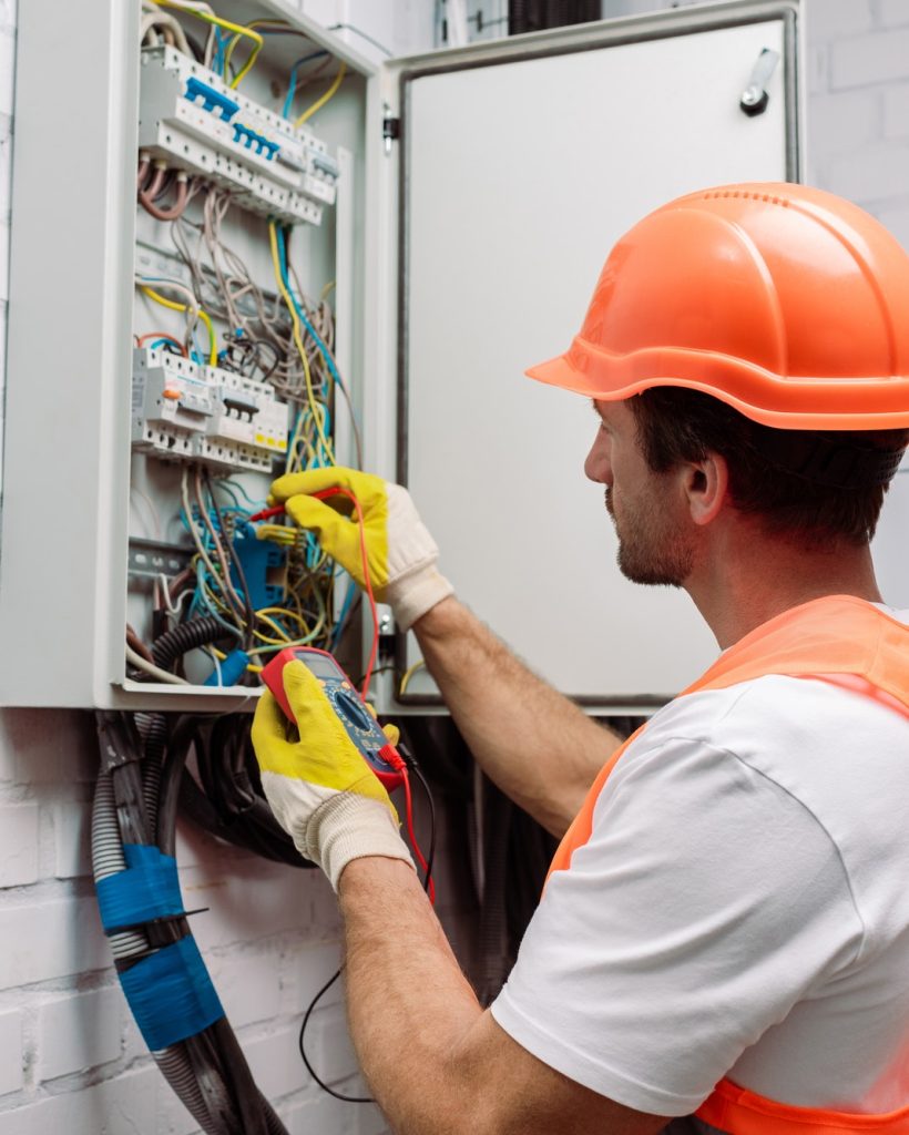 handsome-electrician-in-hardhat-and-gloves-holding-multimeter-near-electrical-distribution-box